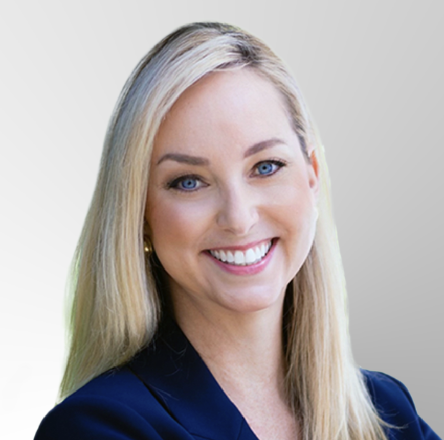 A woman with long blonde hair, wearing a dark blazer, smiling at the camera in front of a light gray gradient background.