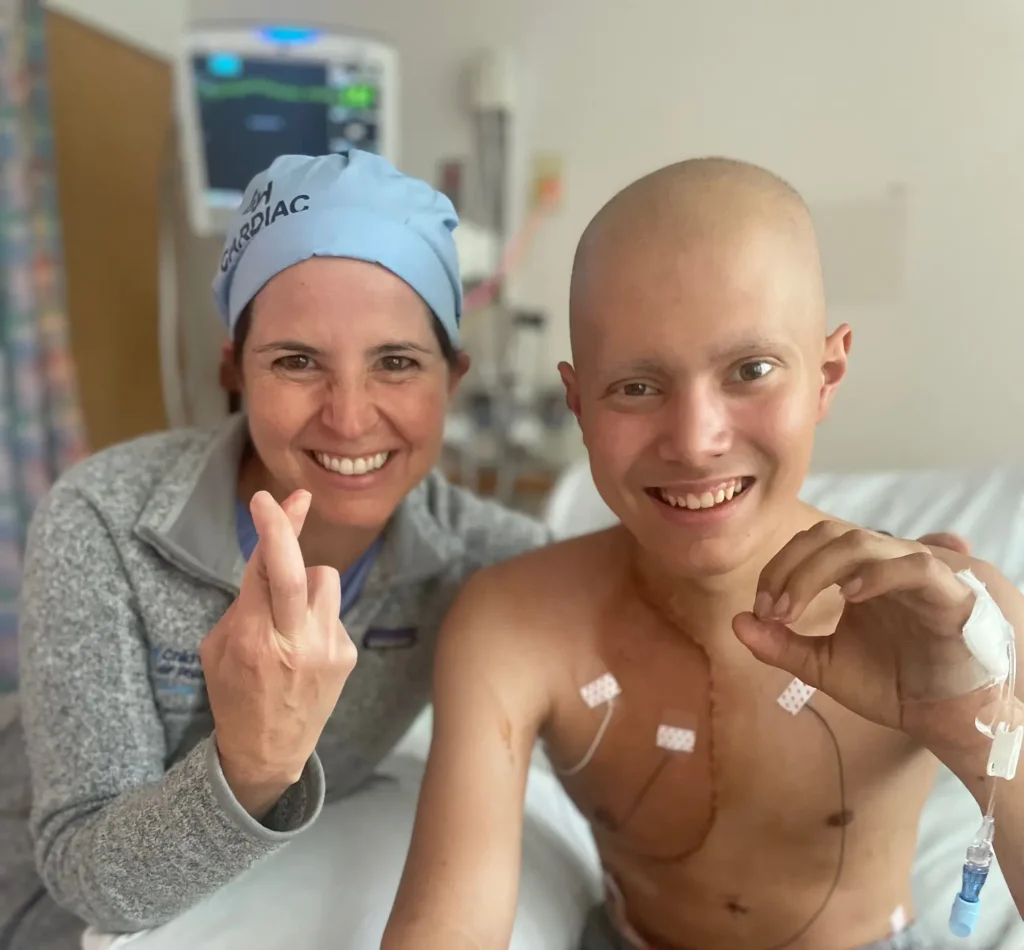 A smiling patient with a bald head and visible chest scars sits on a hospital bed next to a nurse. Both are smiling and holding up crossed fingers, with medical equipment visible in the background.