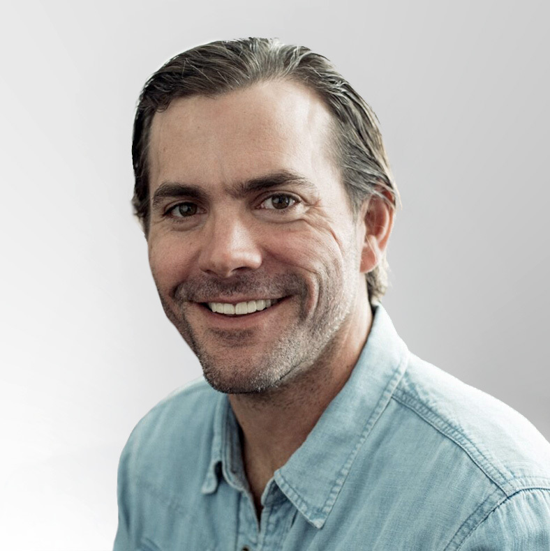 A man with short brown hair and a light beard is smiling. He is wearing a light blue denim shirt and is posed against a plain, light gray background.