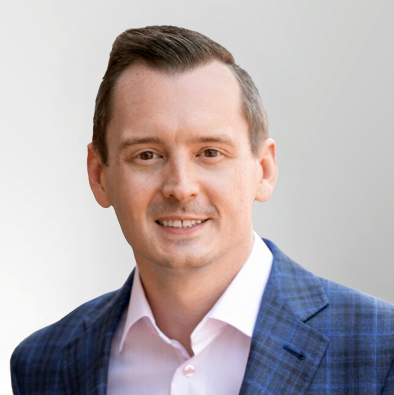 A man with short brown hair wearing a blue plaid suit jacket and a white dress shirt smiles at the camera against a light gray background.