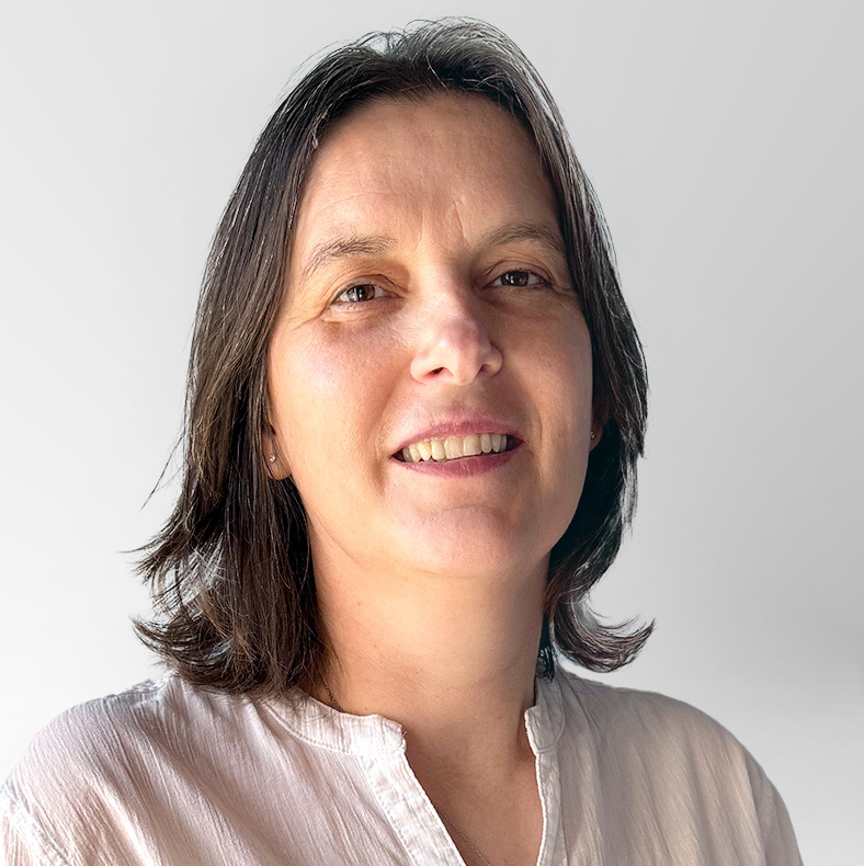 A woman with straight, shoulder-length brown hair and wearing a light-colored blouse smiles gently while looking at the camera against a plain, light gray background.
