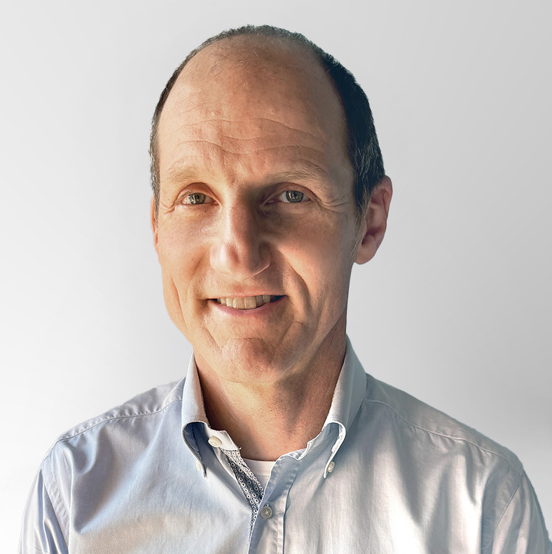 A middle-aged man with short, thinning hair smiles at the camera. He is wearing a light blue button-up shirt and is posed against a plain, light gray background.