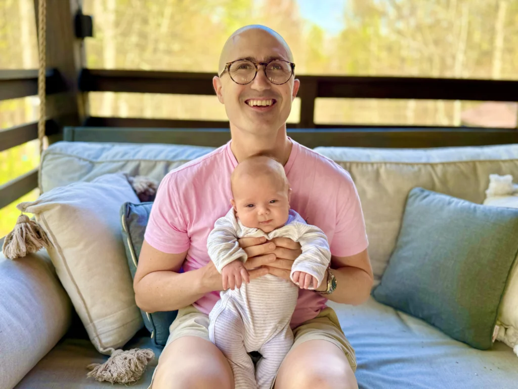 A smiling adult in glasses and a pink shirt sits on an outdoor couch, holding a baby dressed in light-colored clothes on their lap. Trees and greenery are visible in the background.