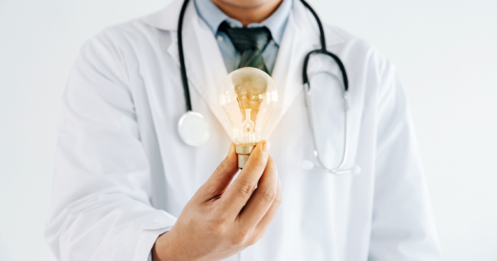 A doctor wearing a white coat and stethoscope holds a glowing light bulb in one hand, symbolizing innovation or new ideas in healthcare.